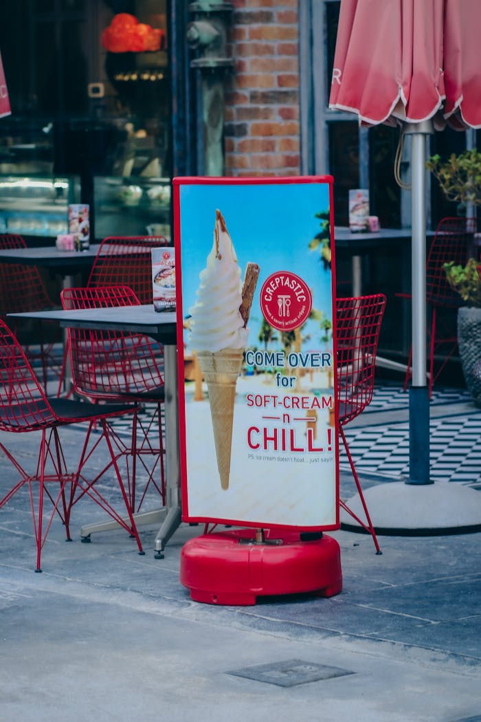 Street view of a cafe with an ice cream advertisement, red seating, and pavement.